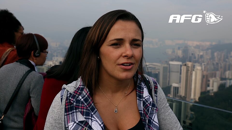 Michelle Nicolini in een geruit shirt tijdens een interview met uitzicht over een stad