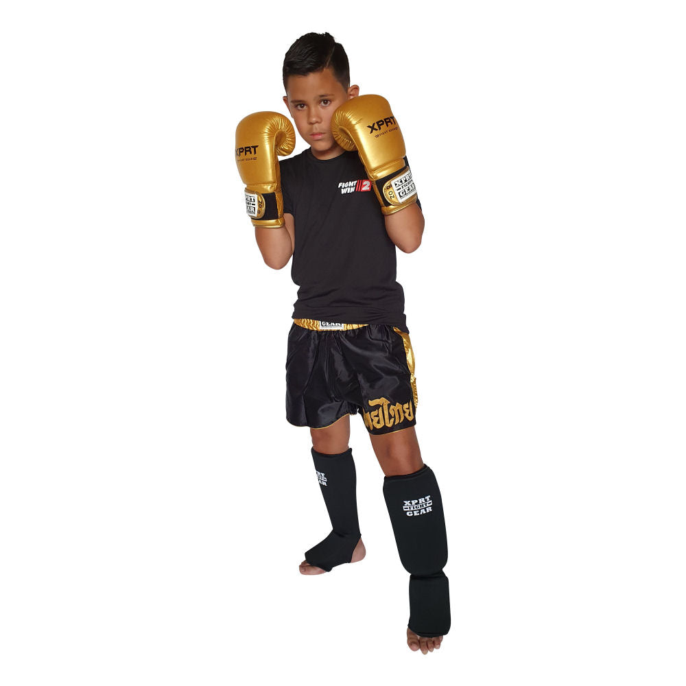 Kind met goudkleurige XPRT-bokshandschoenen, zwarte scheenbeschermers en een zwart-gouden kickboksbroekje.